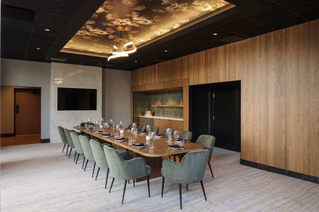 Hilton Arlington Rosslyn The Key Locket Restaurant Chef's Table wooden wall with dry bar with a mini fridge and white countertop backdrop with green tiles and wooden floating shelf next to a marble wall with television mounted in a room with a long wooden table with arm chairs with black legs and green fabric and a recessed ceiling with floral pattern and modern chandelier light fixture