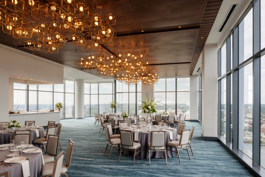 Hilton Arlington Rosslyn The Key The View, a room with floor to ceiling windows with banquet style round tables set for dining with floral arrangements in the center and a bar and abstract cluster of cubes light fixture