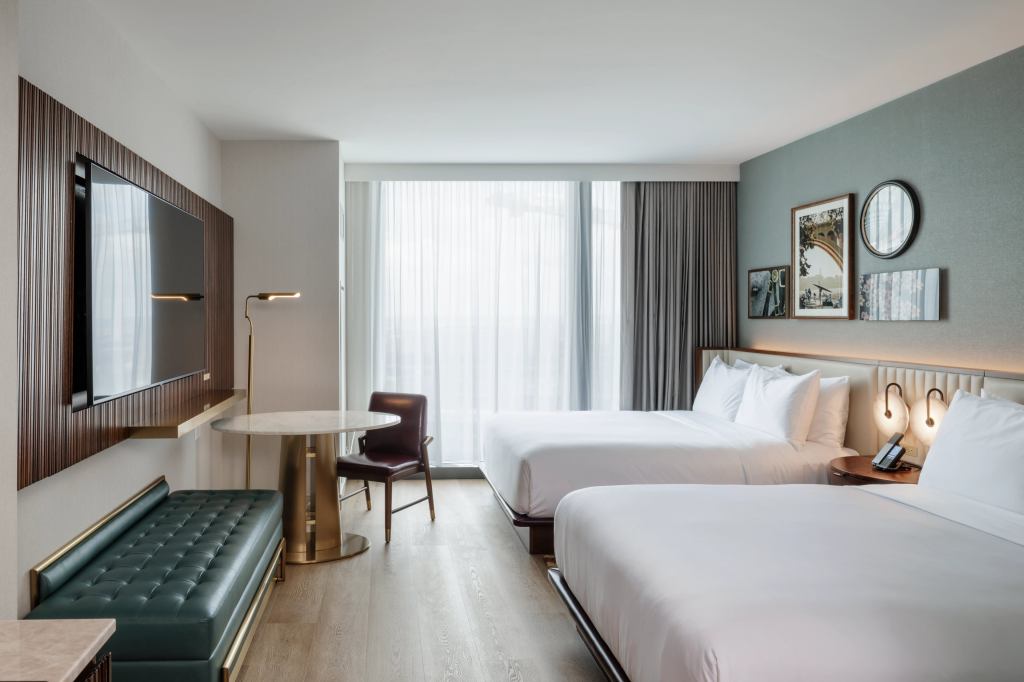 Hilton Arlington Rosslyn The Key Two Queen Guest Room with beds with white bedding and an end table in between with mounted round light fixtures and a telephone with framed pictures on wall and a window next to a wall with a accent chair with a round accent table with a marble top and a green upholstered bench with a floor lamp and a television mounted on the wall