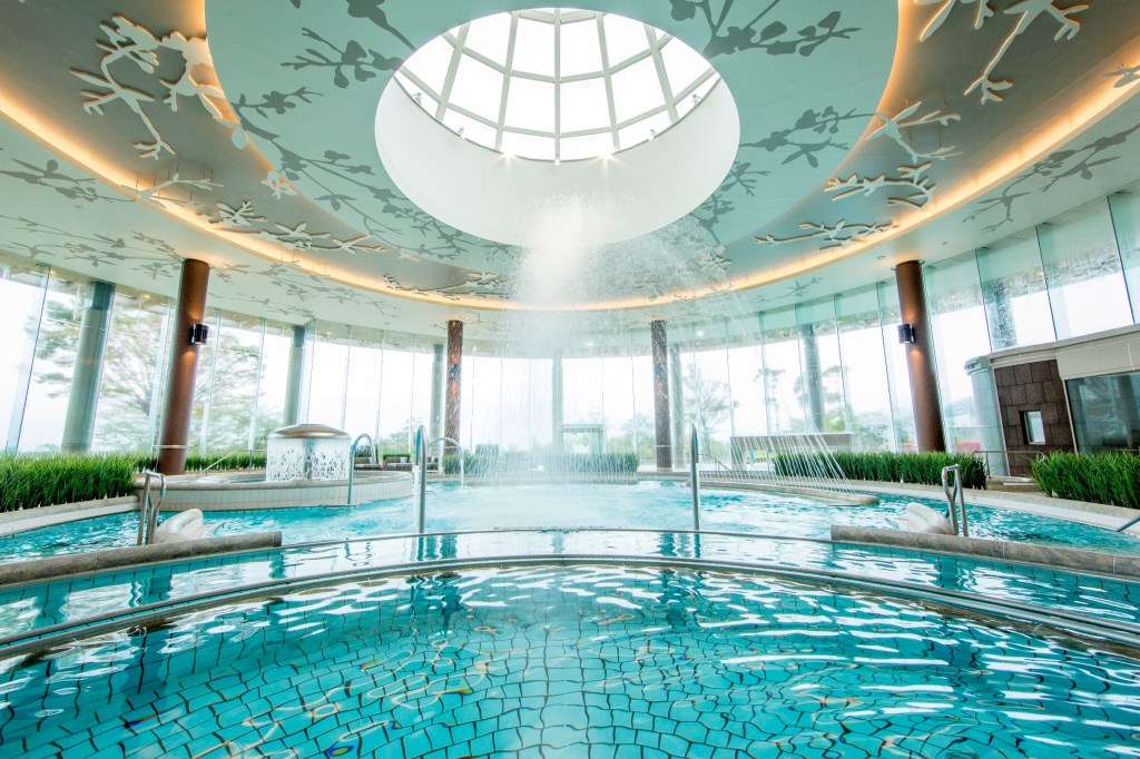 Hilton Odawara Resort &amp; Spa, Bade Zone surrounded by natural lighting from a full wall of exterior windows and skylight above, water fountain, potted plants, tiled pool, artwork on the ceiling with skylight