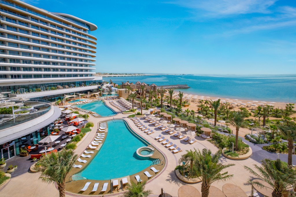 Waldorf Astoria Doha Lusail Pool with lounge seating and umbrellas next to the pool as well as on the beach with palm trees