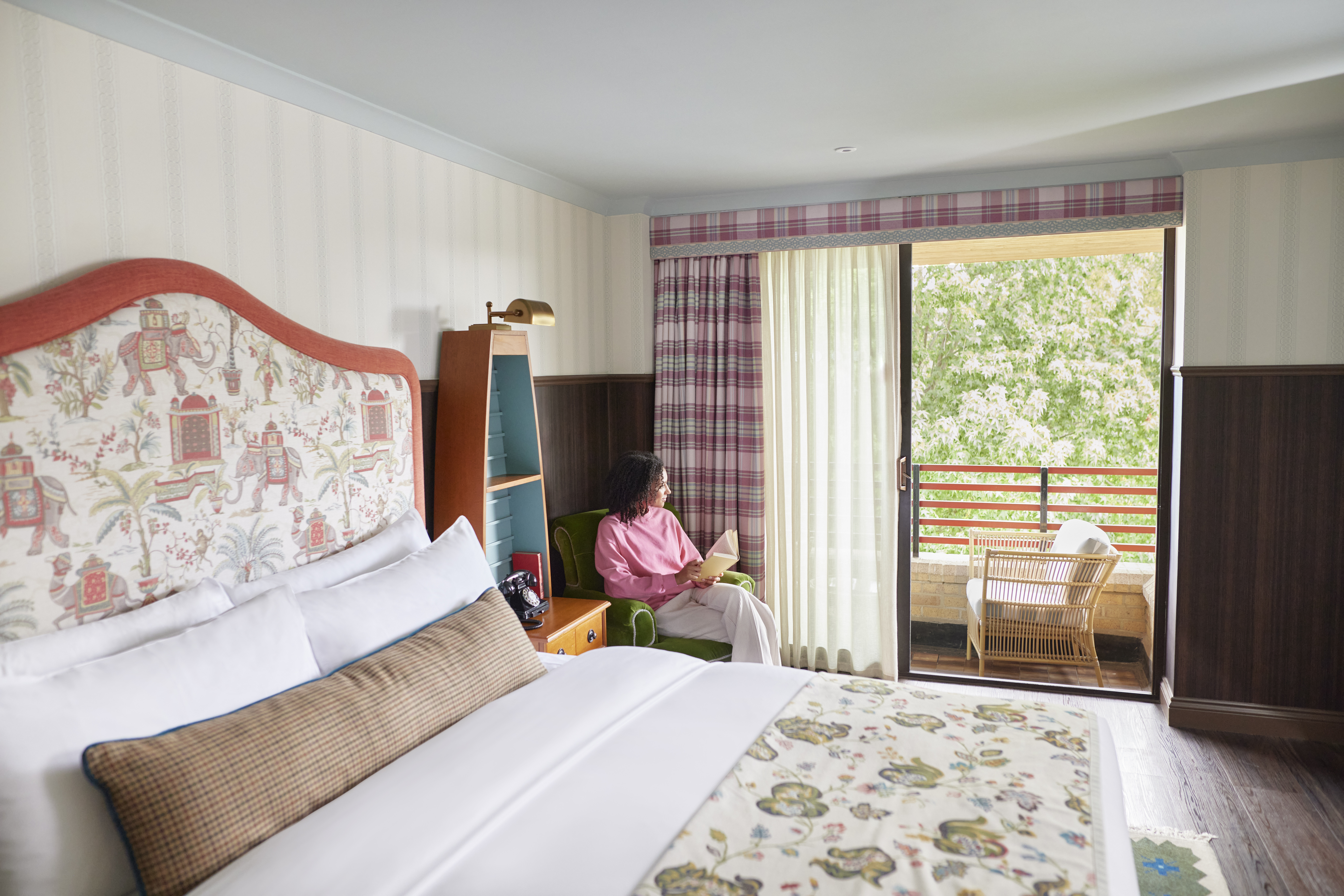 Graduate by Hilton Cambridge - Guest Room, view of bed and balcony with chairs and view of greenery, woman sitting in chair holding a book looking out balcony doors