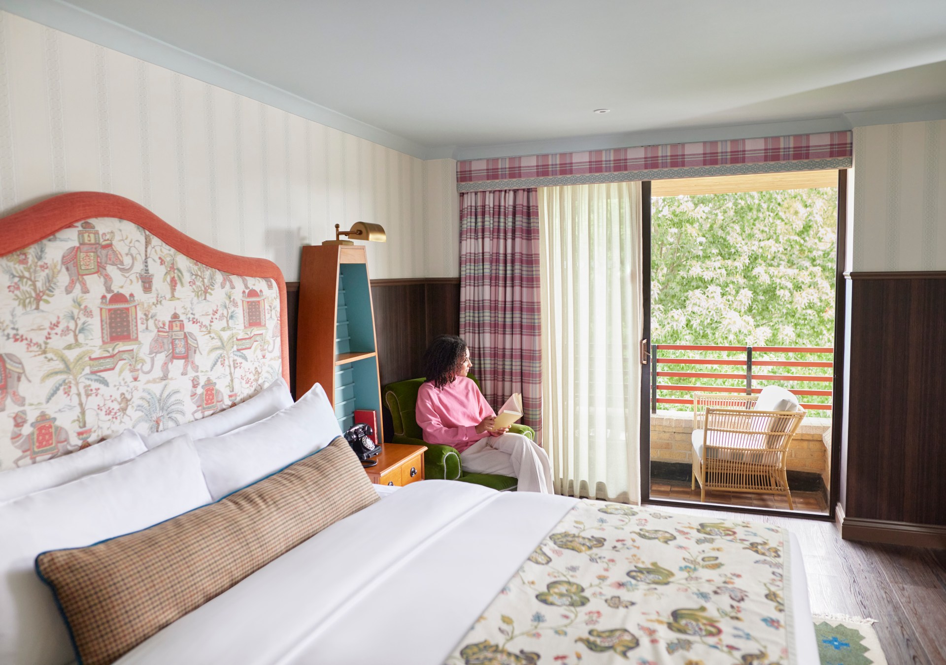 Graduate by Hilton Cambridge - Guest Room, view of bed and balcony with chairs and view of greenery, woman sitting in chair holding a book looking out balcony doors