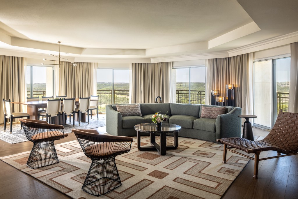 A seating area with a large couch, two chairs, a chaise, and a coffee table with dining table and balcony overlooking the landscape in the background.