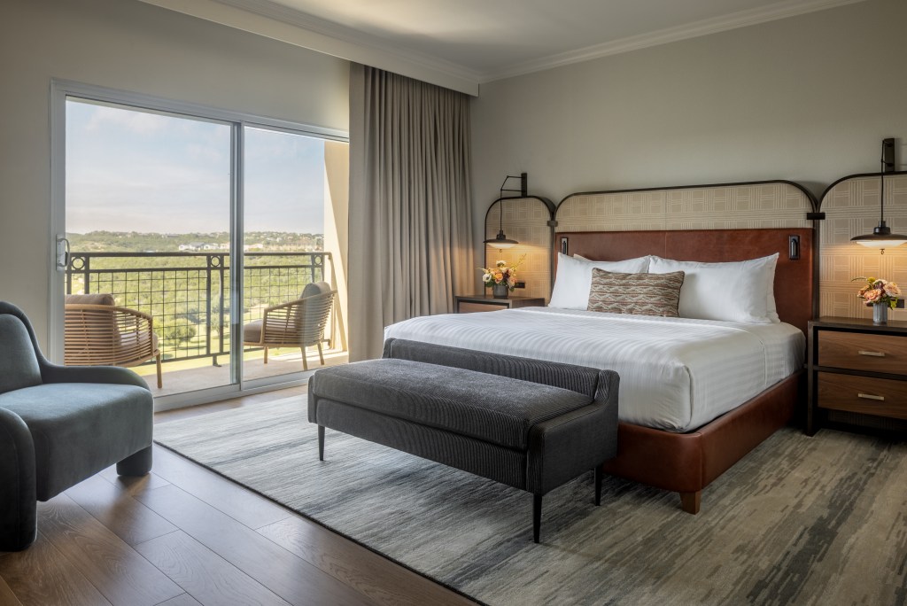 A bedroom with a king bed and chair and a balcony overlooking the landscape