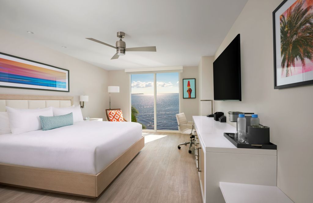 A bedroom with a king bed, tv, desk and chair. There is a large window overlooking the ocean in the background.