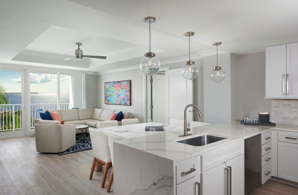 A large room with a kitchen in the foreground, a dining table, and living room area with couch in the background. A large window overlooking the balcony and the ocean fills the back wall.