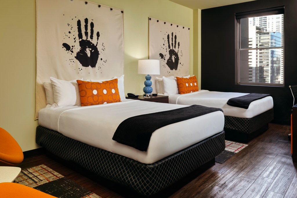 ACME Hotel Chicago - Guest Room - guest room with two beds, handprint artwork above beds, orange pillows on beds, window looking out to the city