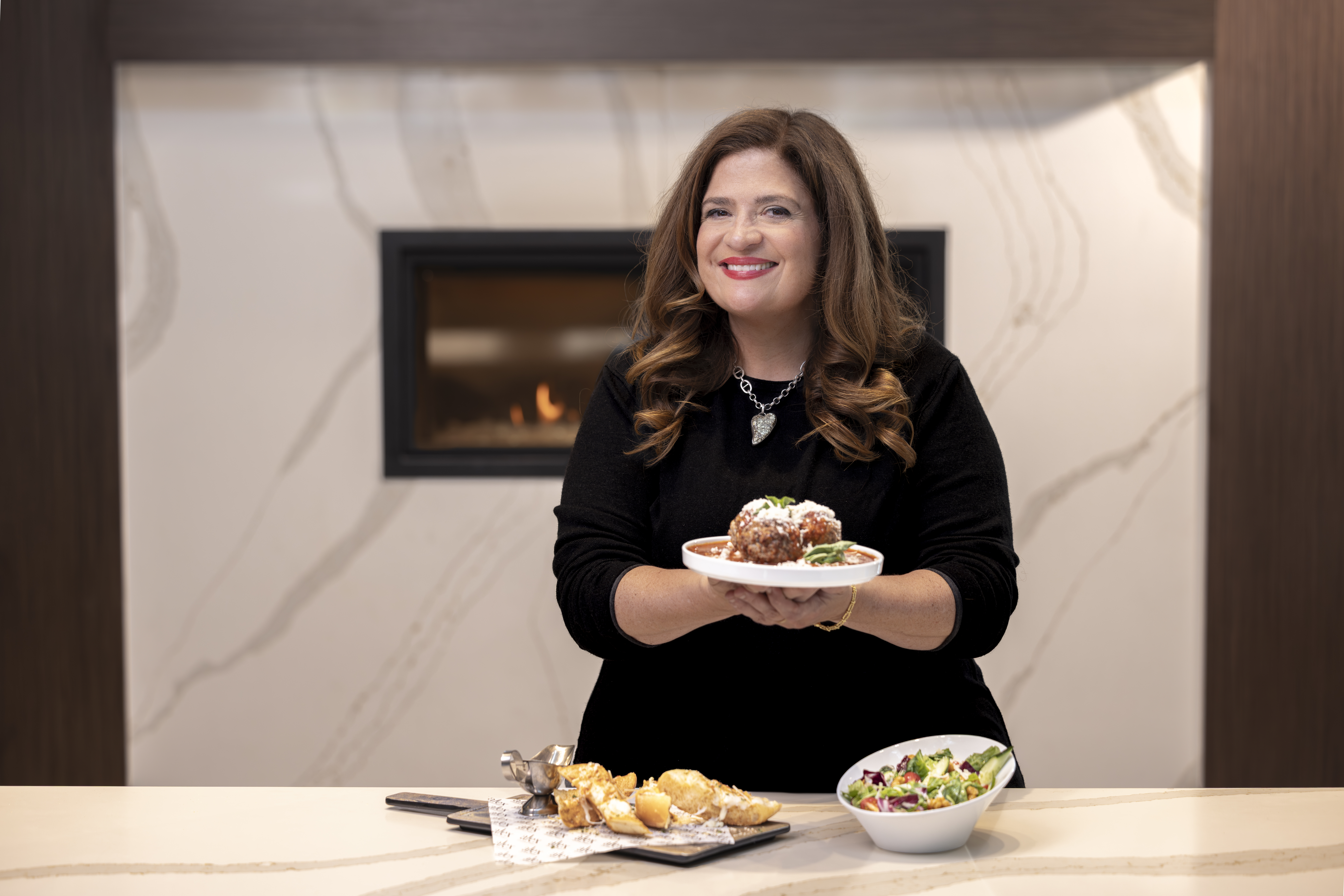 Chef Alex Guarnaschelli holding food, with food on the table in front of her, smiling at camera