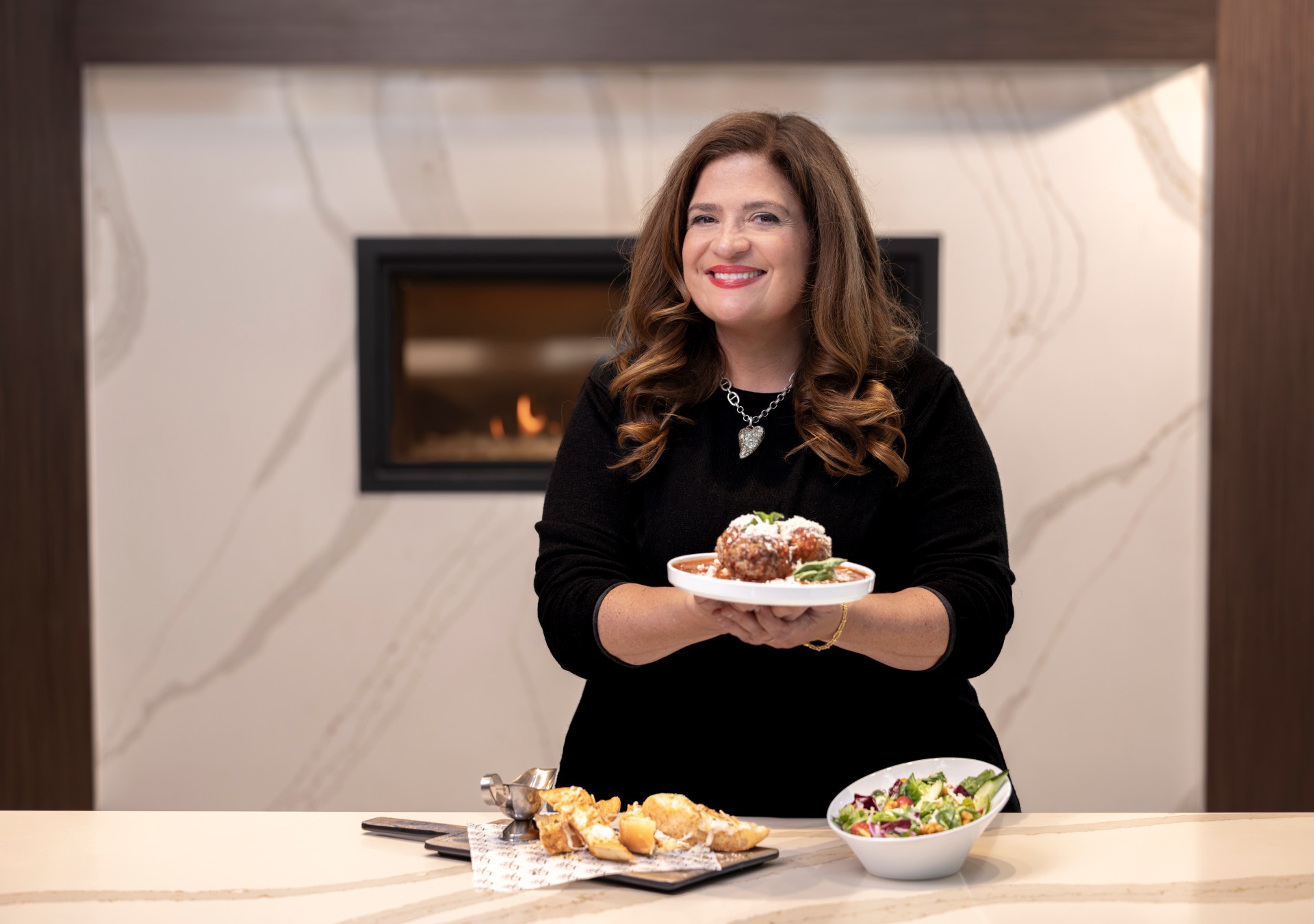 Chef Alex Guarnaschelli holding food, with food on the table in front of her, smiling at camera