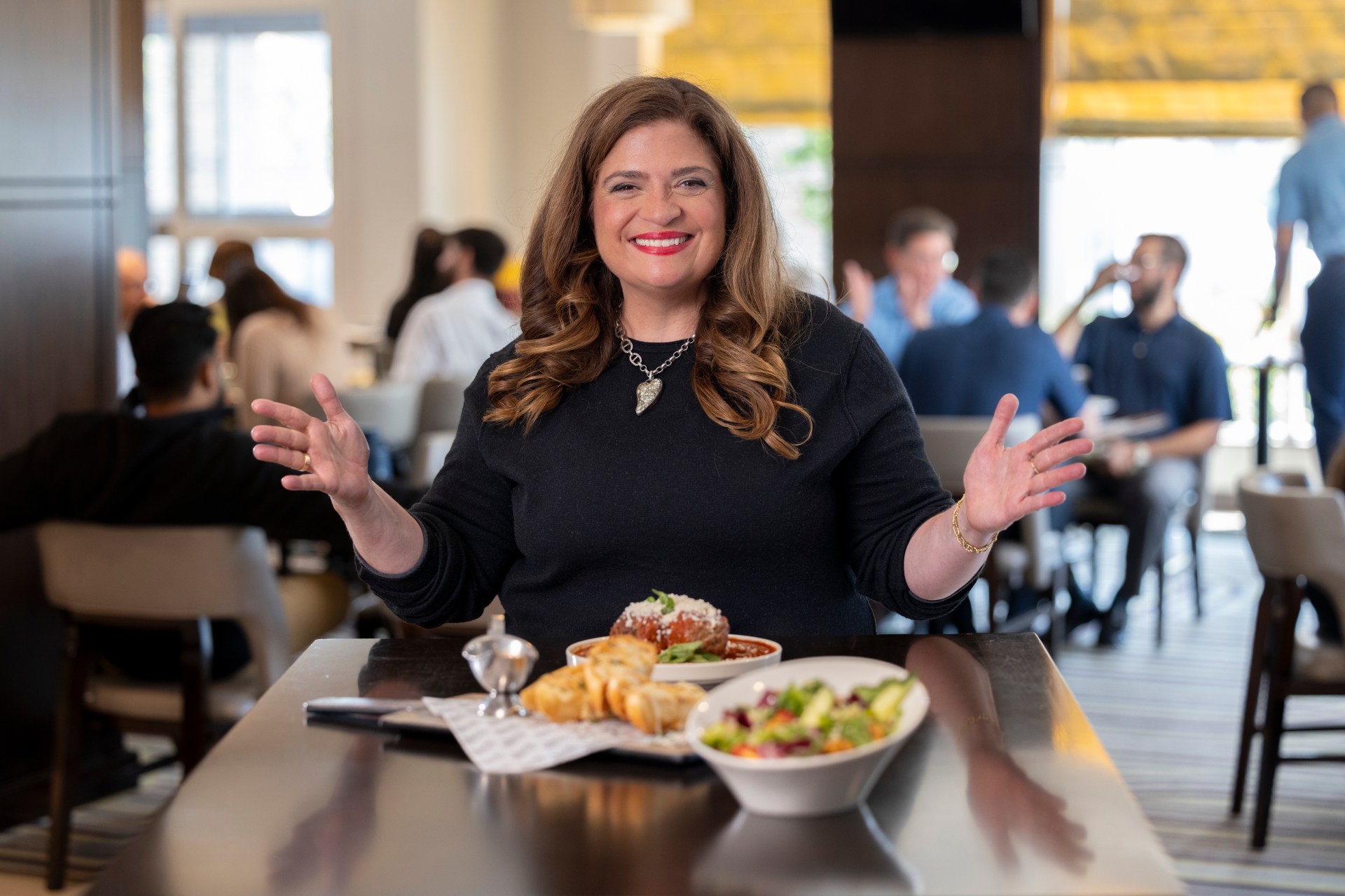 Chef Alex Guarnaschelli in a dining room with guest around her, and her food in front of her, smiling at camera