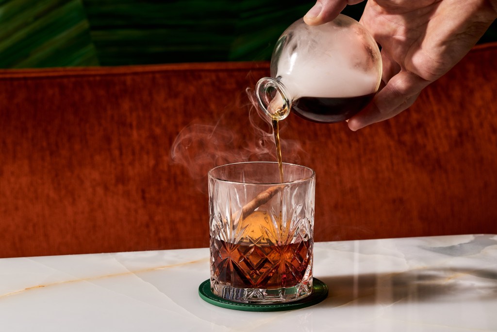 A specialty cocktail being poured from a small bottle, with smoke coming out