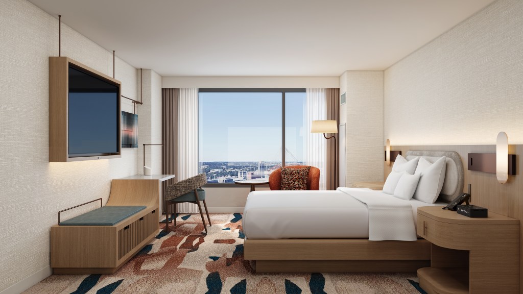 A bedroom with bed, chair, desk, tv, and window looking out at the city
