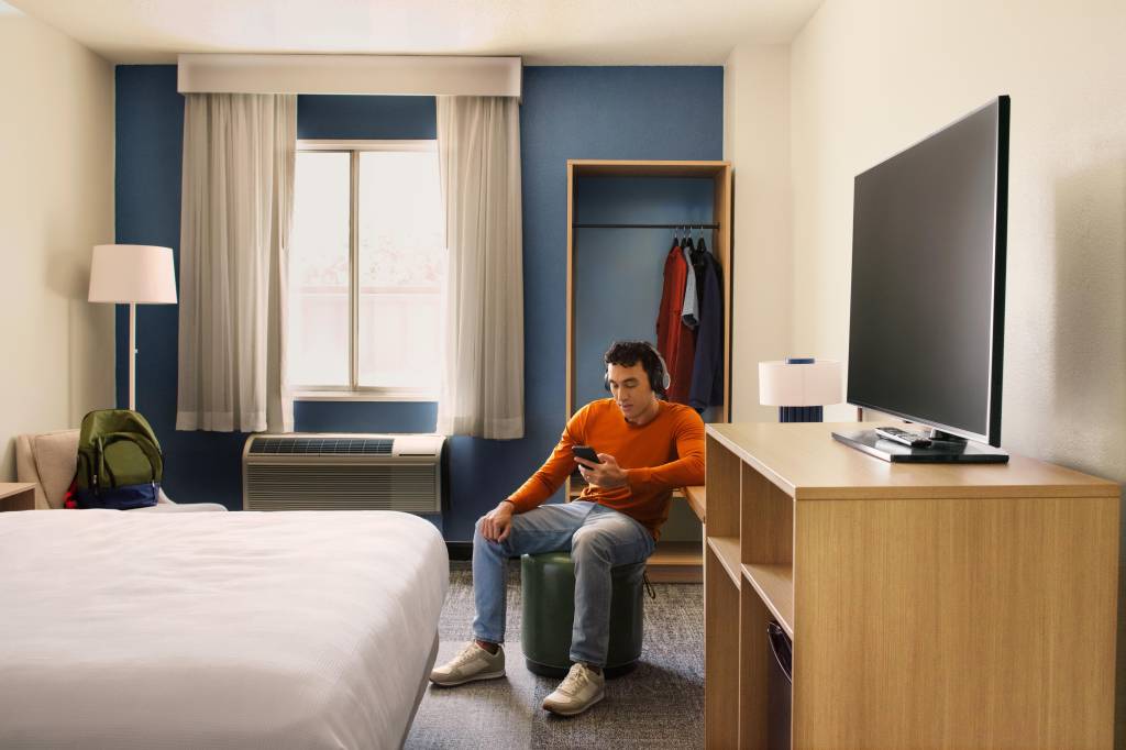 man sitting on a stool wearing headphones and looking at phone in Spark by Hilton guest room with bed, television, closet, chair, window