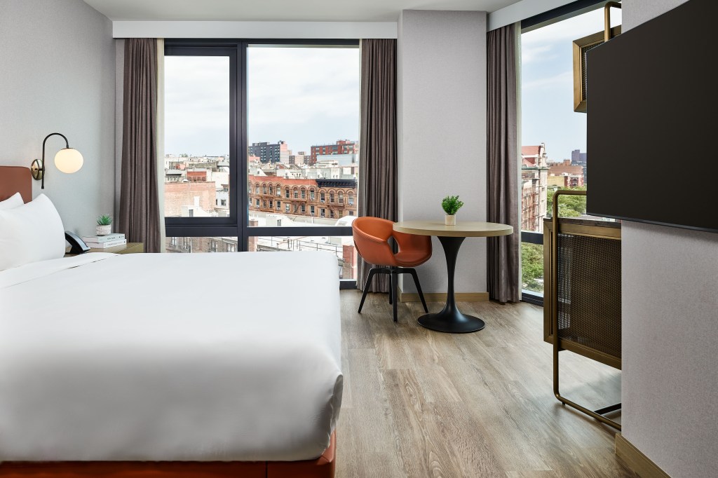 A bright bed room with bed, table and chair, tv, and windows looking out at the city.