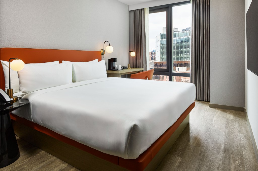 A bedroom with bed, desk, and window looking out at the city