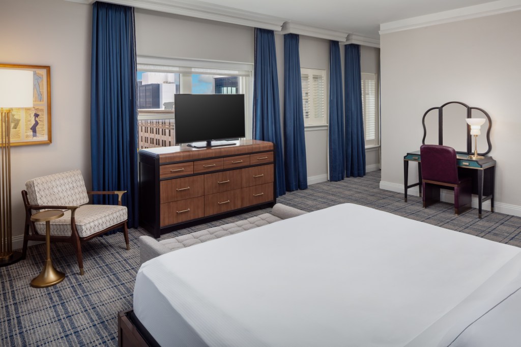 A bedroom with bed, bench, chair, vanity table, and tv