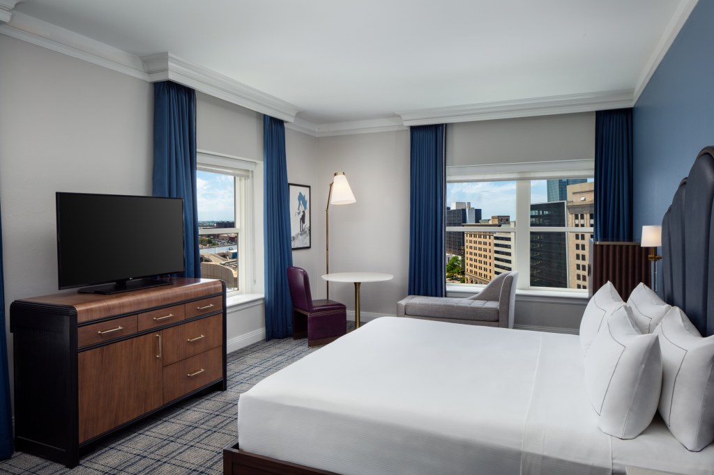 A room with bed, tv, chair and table, and chaise in front of windows overlooking the city