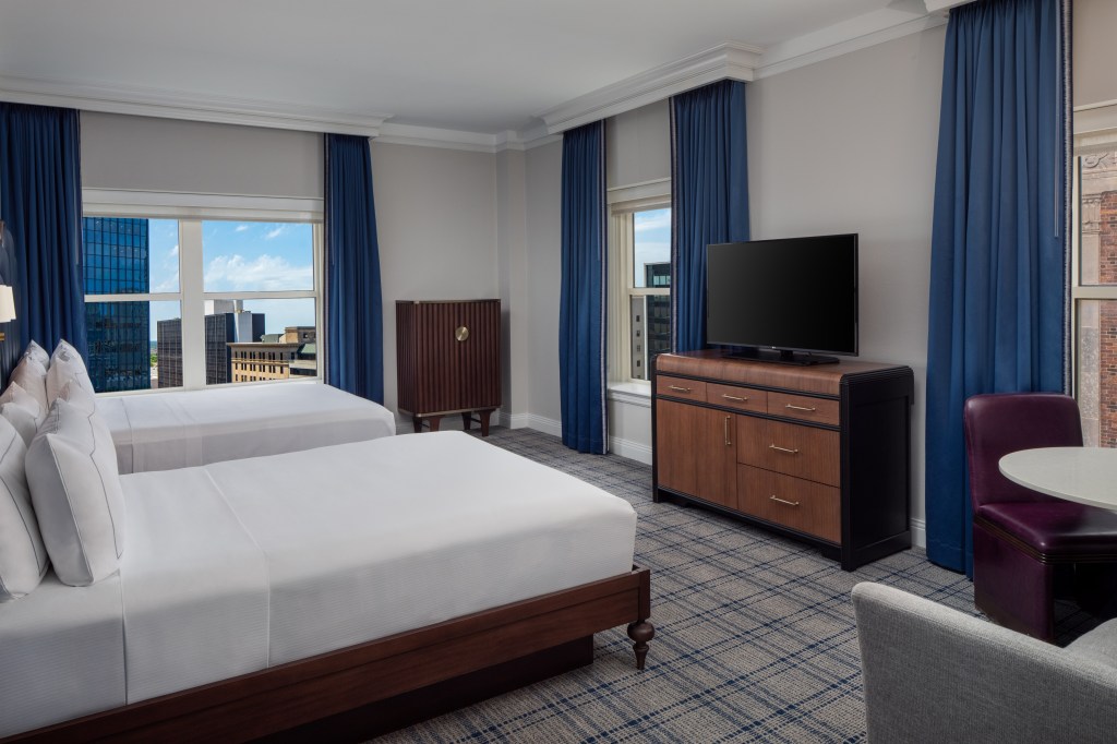 A bed room with two beds, tv, table and chairs, and windows overlooking the city.