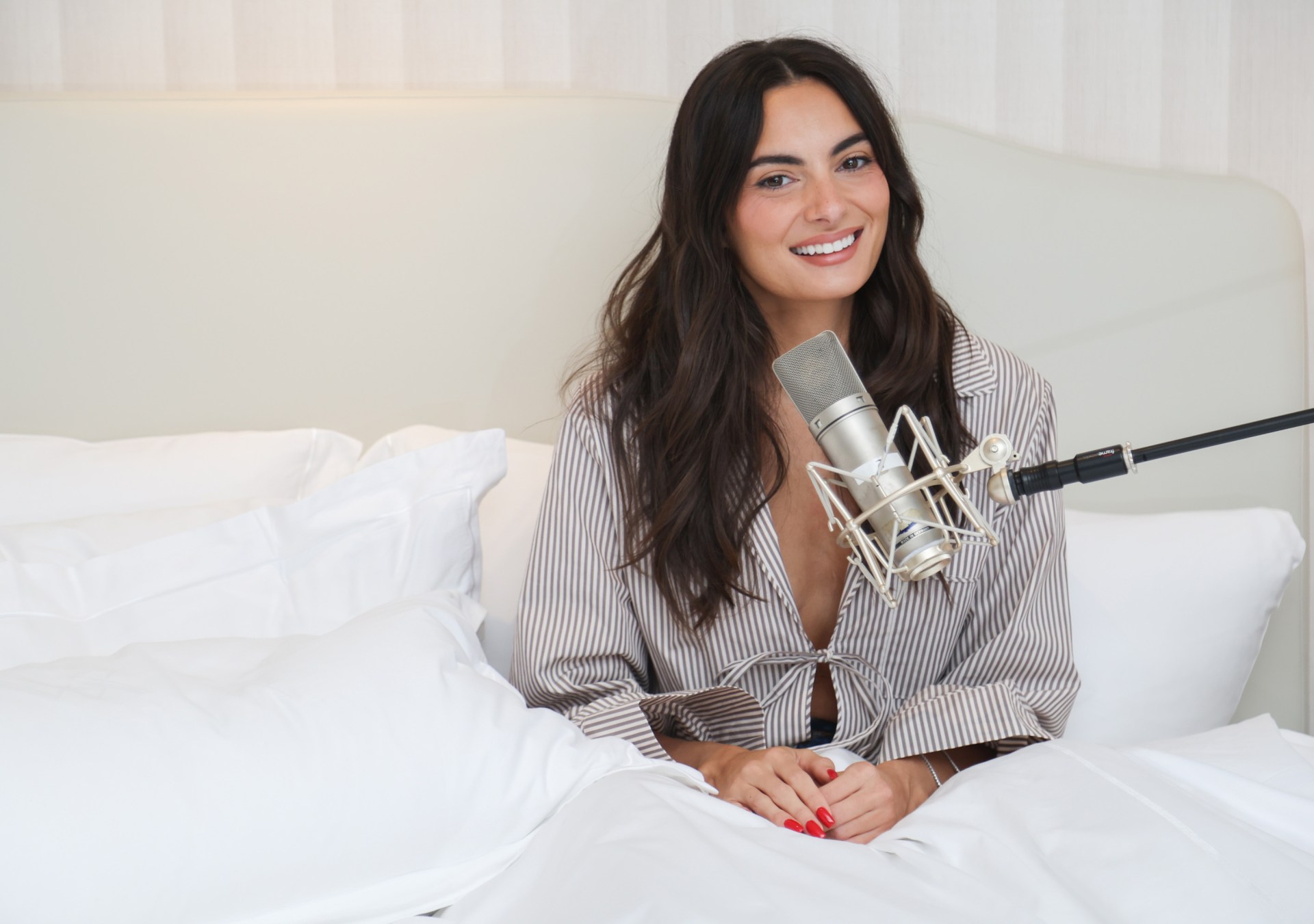 Paige DeSorbo sits in bed with microphone, smiling at camera