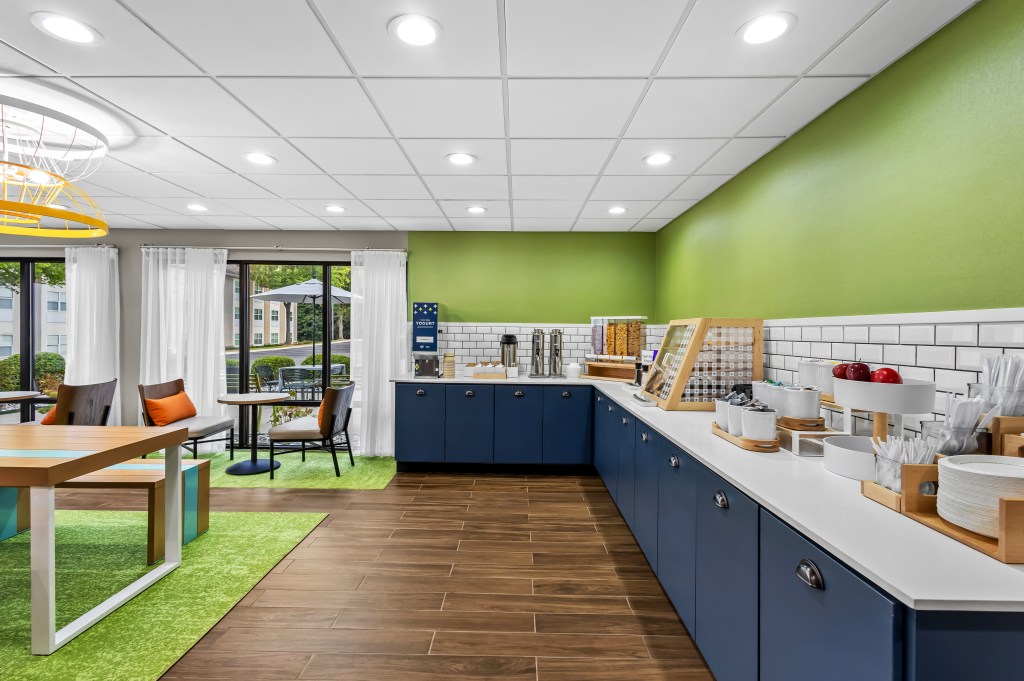 Spark by Hilton Alpharetta Roswell breakfast bar with lounge seating area and table seating area with bench, apples in a bowl, utensil organizer, pastry display, cereal dispenser