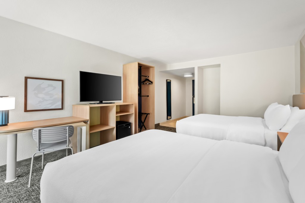 Spark by Hilton Chattanooga Downtown guest room with two beds, desk with grey chair, framed white board, television, tv, mini fridge, mirror, wooden wardrobe, wooden armoire, white bedding, blue desk lamp