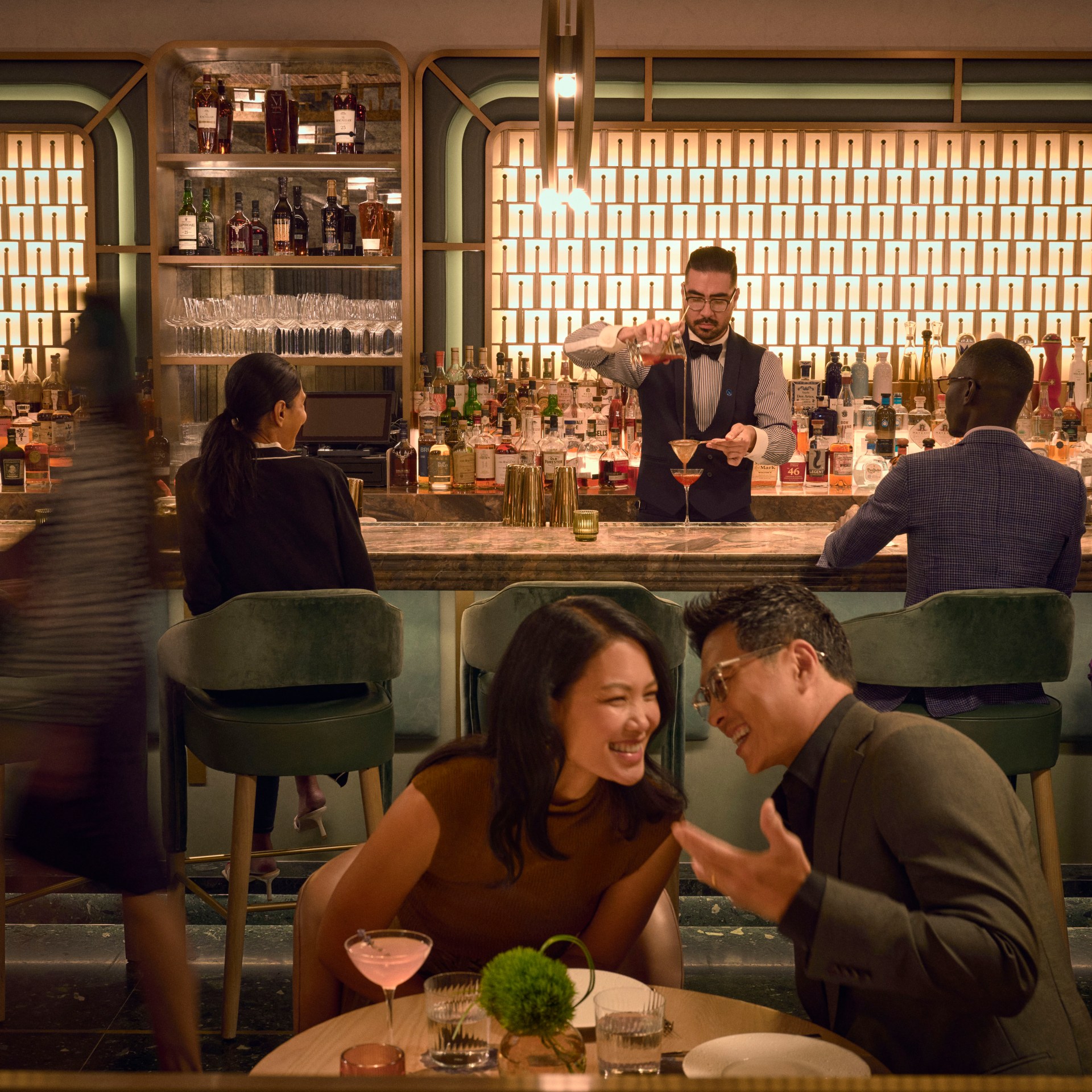 Waldorf Astoria New York - Lex Yard, People sitting on green upholstered barstools and leather chairs in a bar drinking cocktails in coup glassed with a bartender pouring a drink, American brasserie, casually elegant dining room and lively bar, green bar stools
