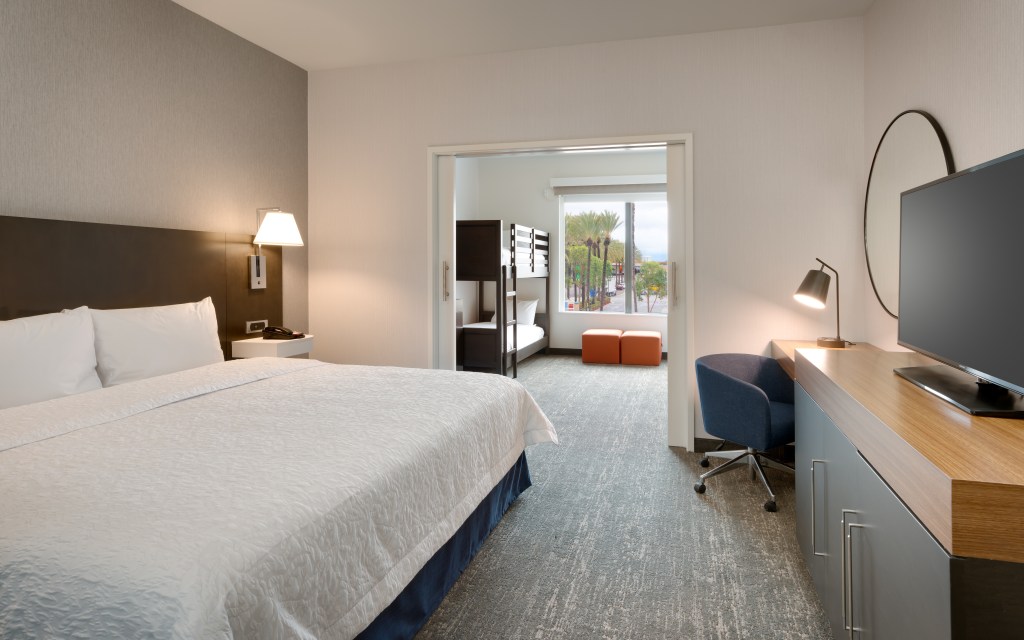 Hampton Inn & Suites Anaheim Resort Convention Center - King bed, television, desk, chair, open pocket door leading out to room with bunk beds, window, orange seating area