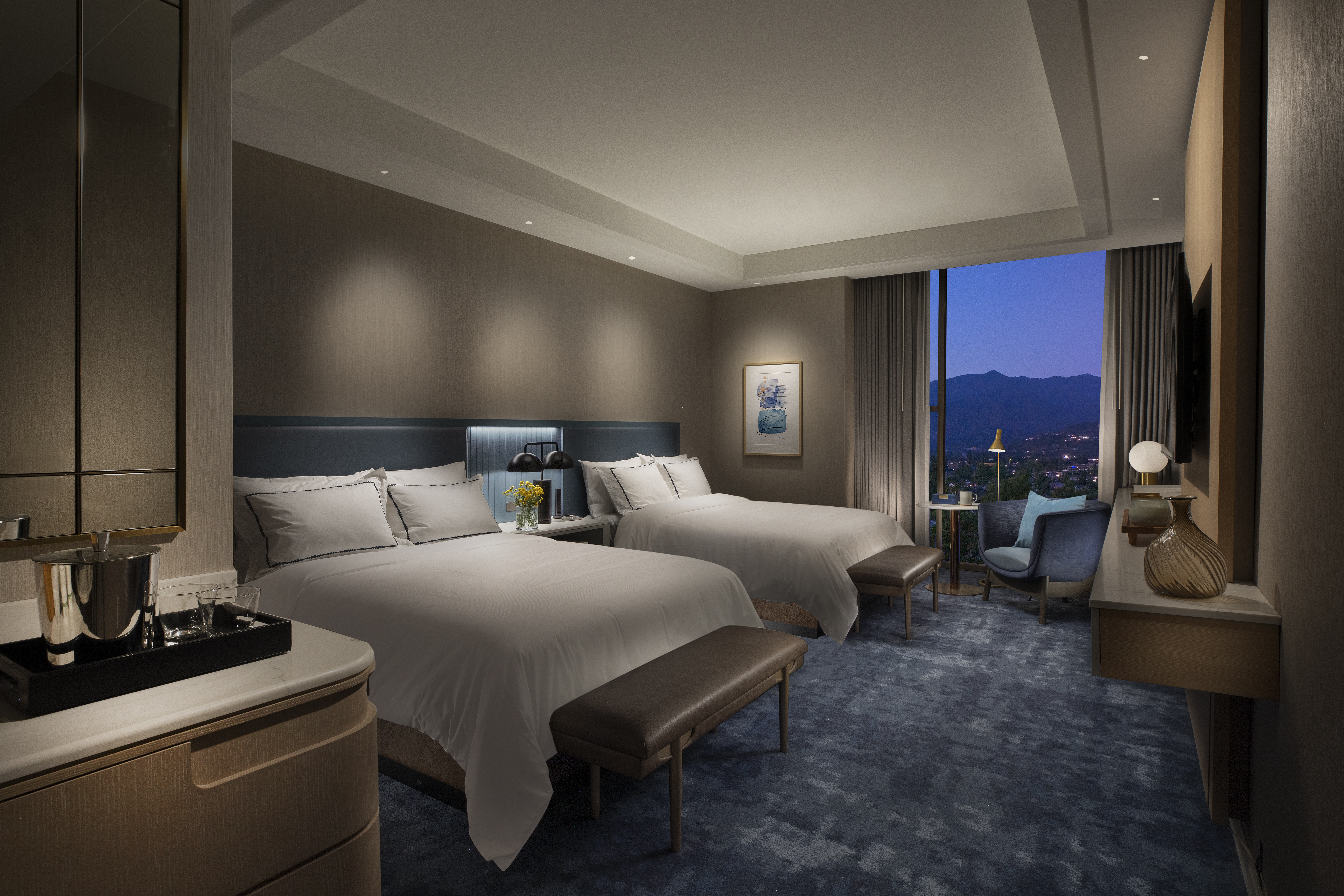 A room with two beds, table and chair, and window overlooking the city