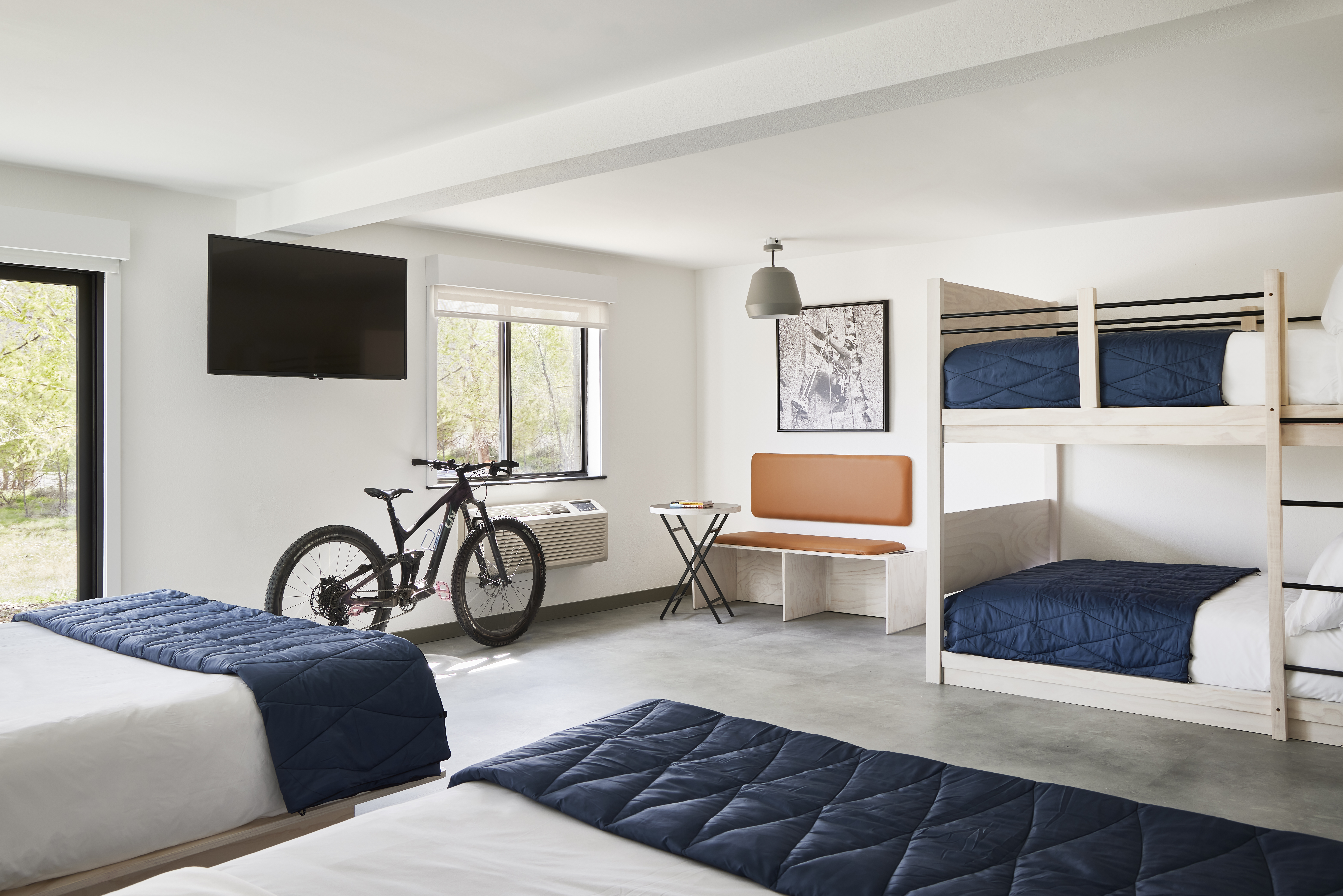A room with two regular beds, a set of bunk beds, a bench, table, tv, and bike