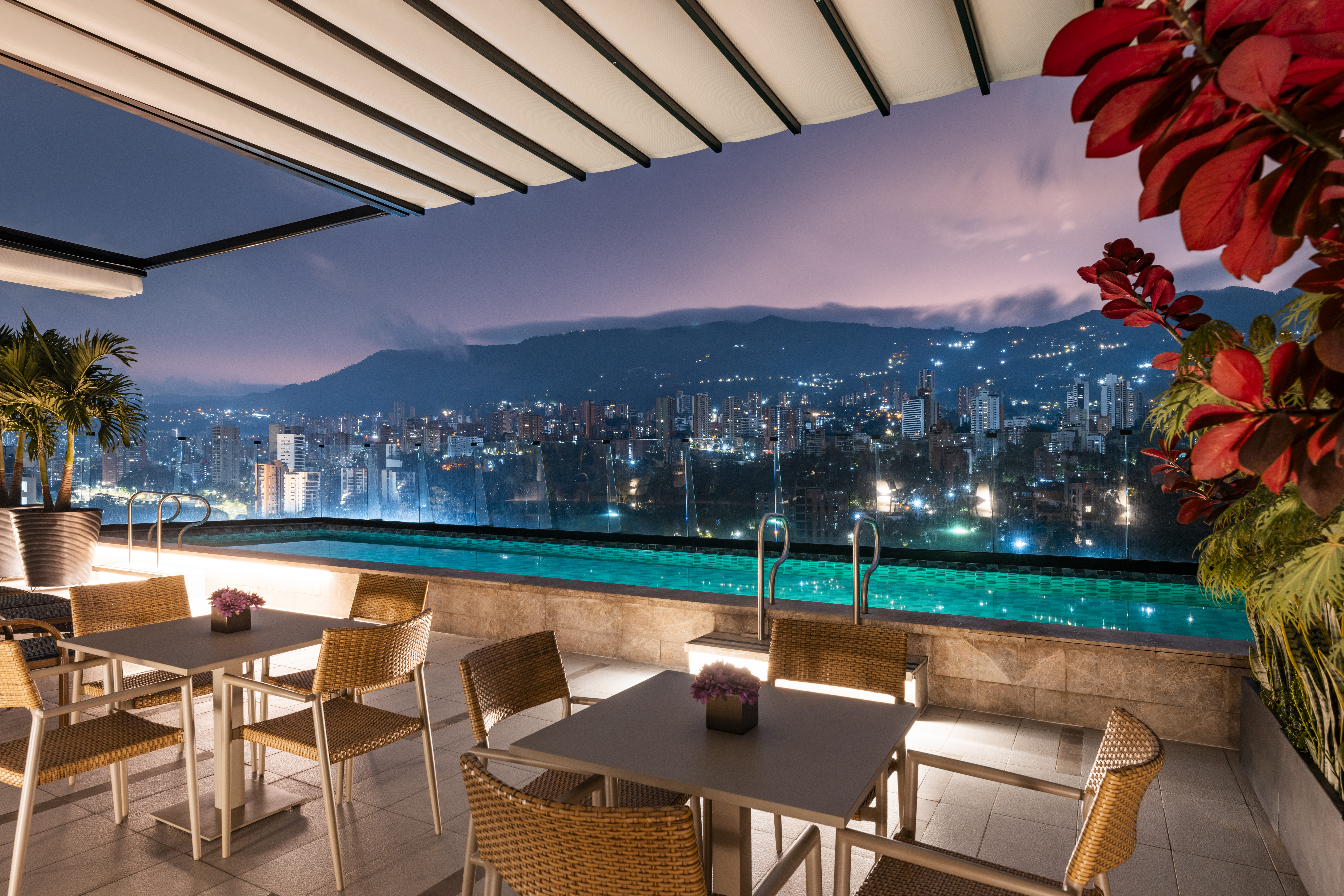 tables and chairs next to the rooftop pool at night, with the lights of the city in the background