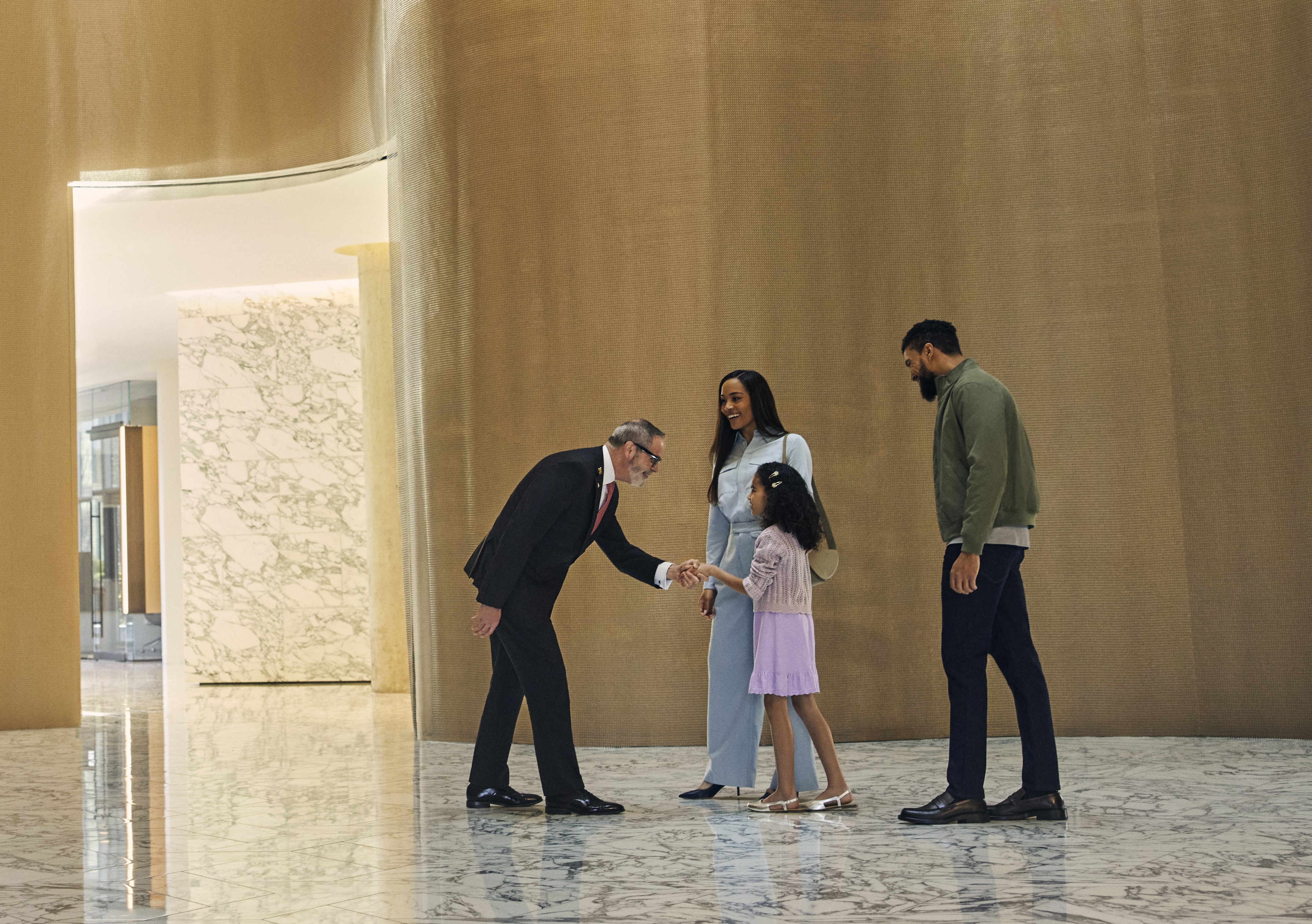 Hilton team member in suit shaking hands with young girl in a modern lobby, with smiling adults nearby and marble floors in a bright interior.