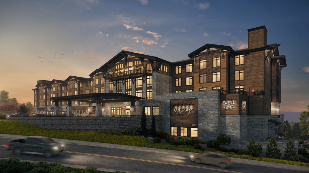 Modern rustic hotel with wood and stone exterior, lit windows, covered entrance, balconies, and Canopy by Hilton signage at dusk.
