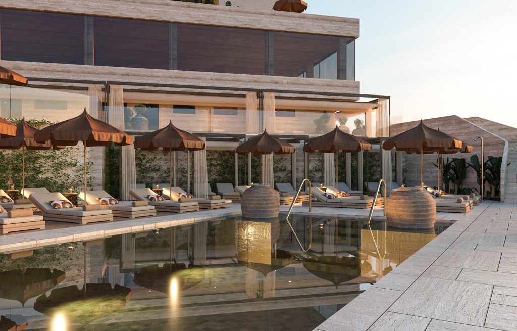 Modern outdoor pool with stone deck, cushioned lounge chairs, brown umbrellas, and glass-front building with greenery in a calm setting.