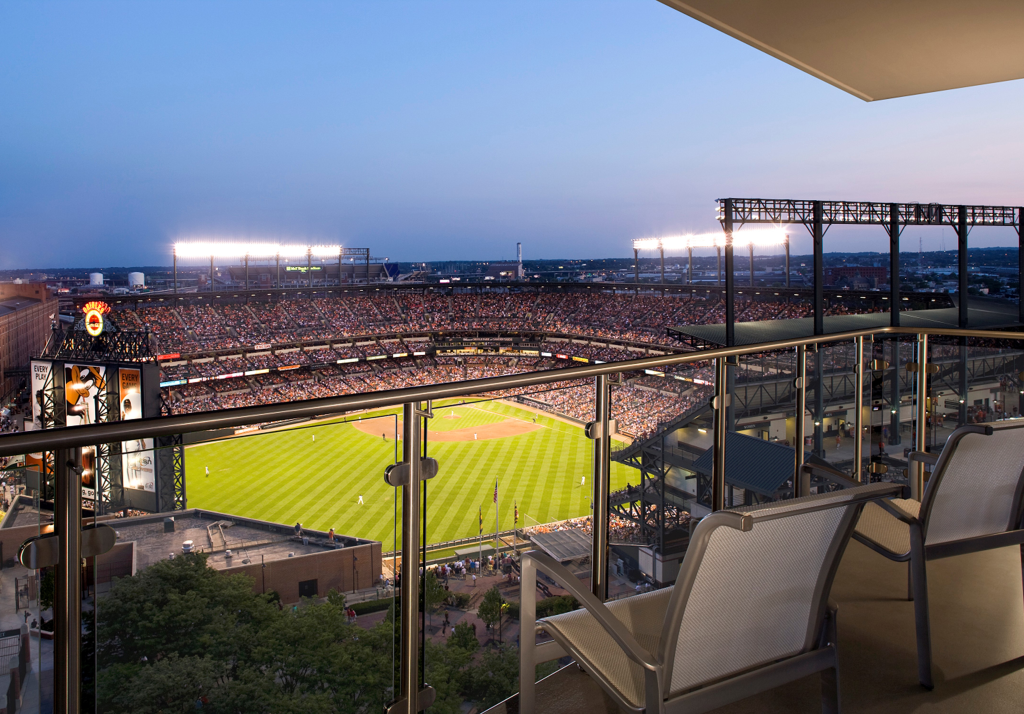Balcony view of a lit baseball stadium at dusk, with empty chairs, glass railing, green field, and packed stands under a purple-blue sky.