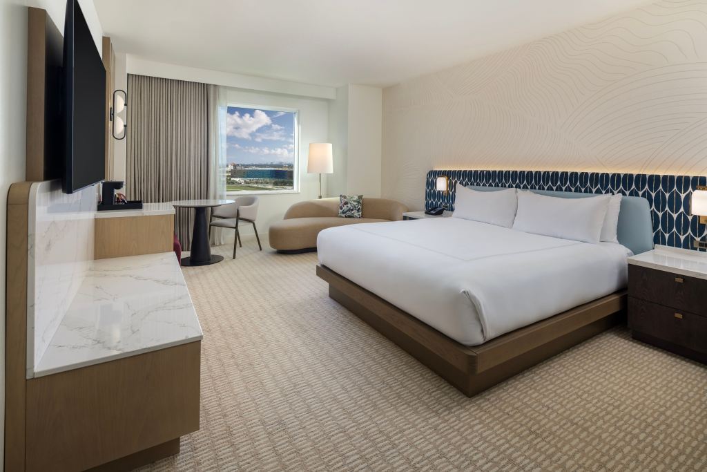 Modern hotel room with king bed, blue headboard, sofa seating, desk by window, TV, and city views in a bright, contemporary setting.