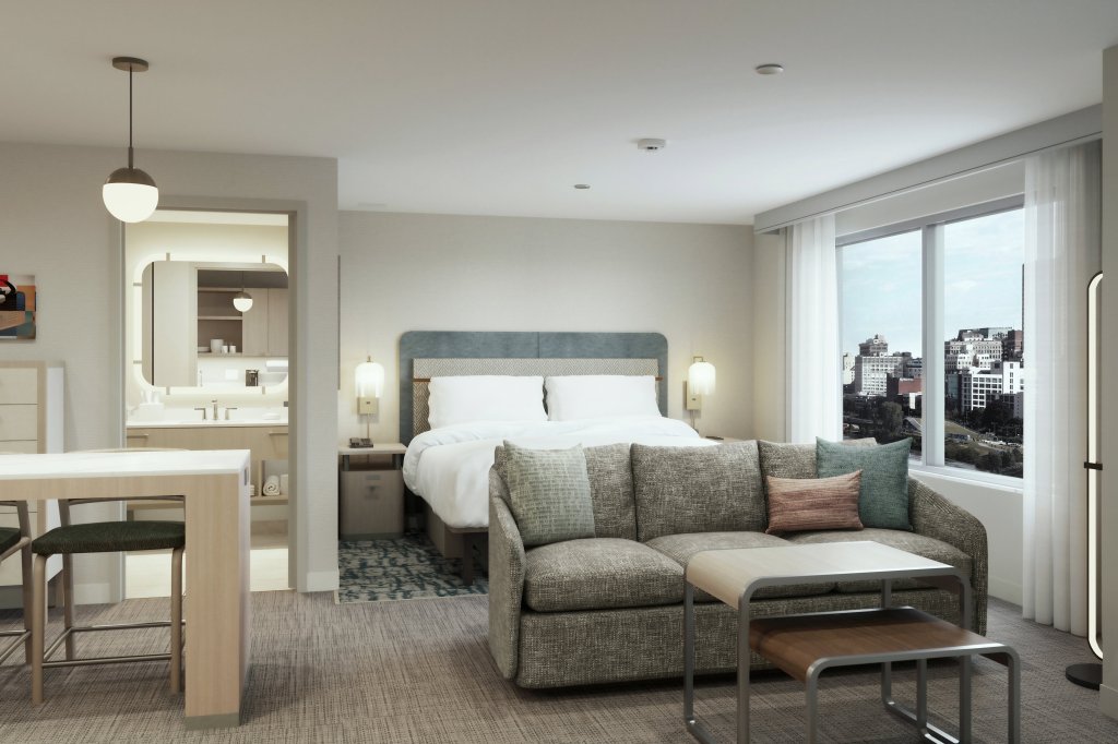 Modern hotel room with blue headboard bed, sofa seating, coffee tables, dining area, city-view window, and open bathroom with illuminated mirror.