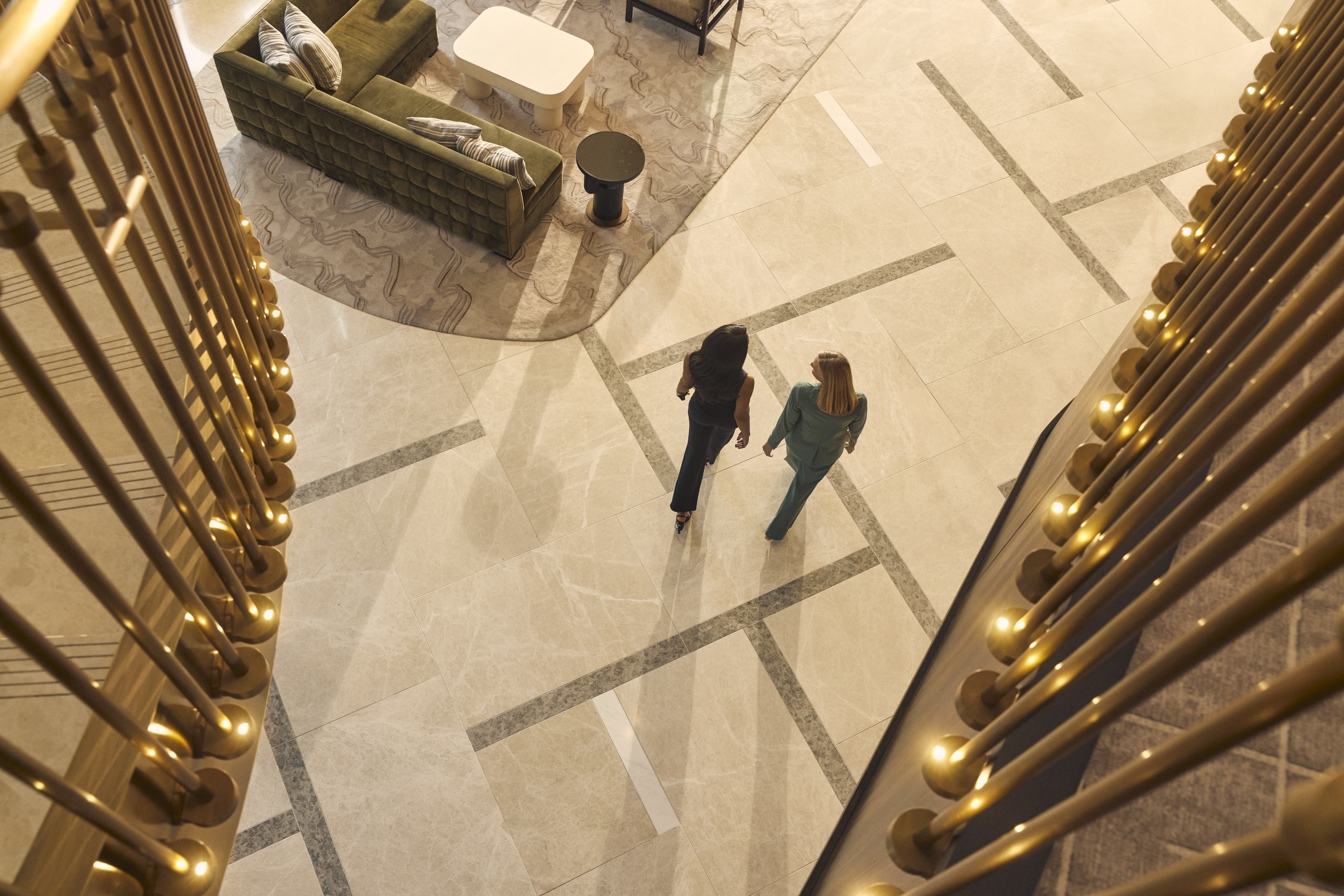 Two women walking on a tiled floor in a modern lobby with a green sofa and golden railings visible.