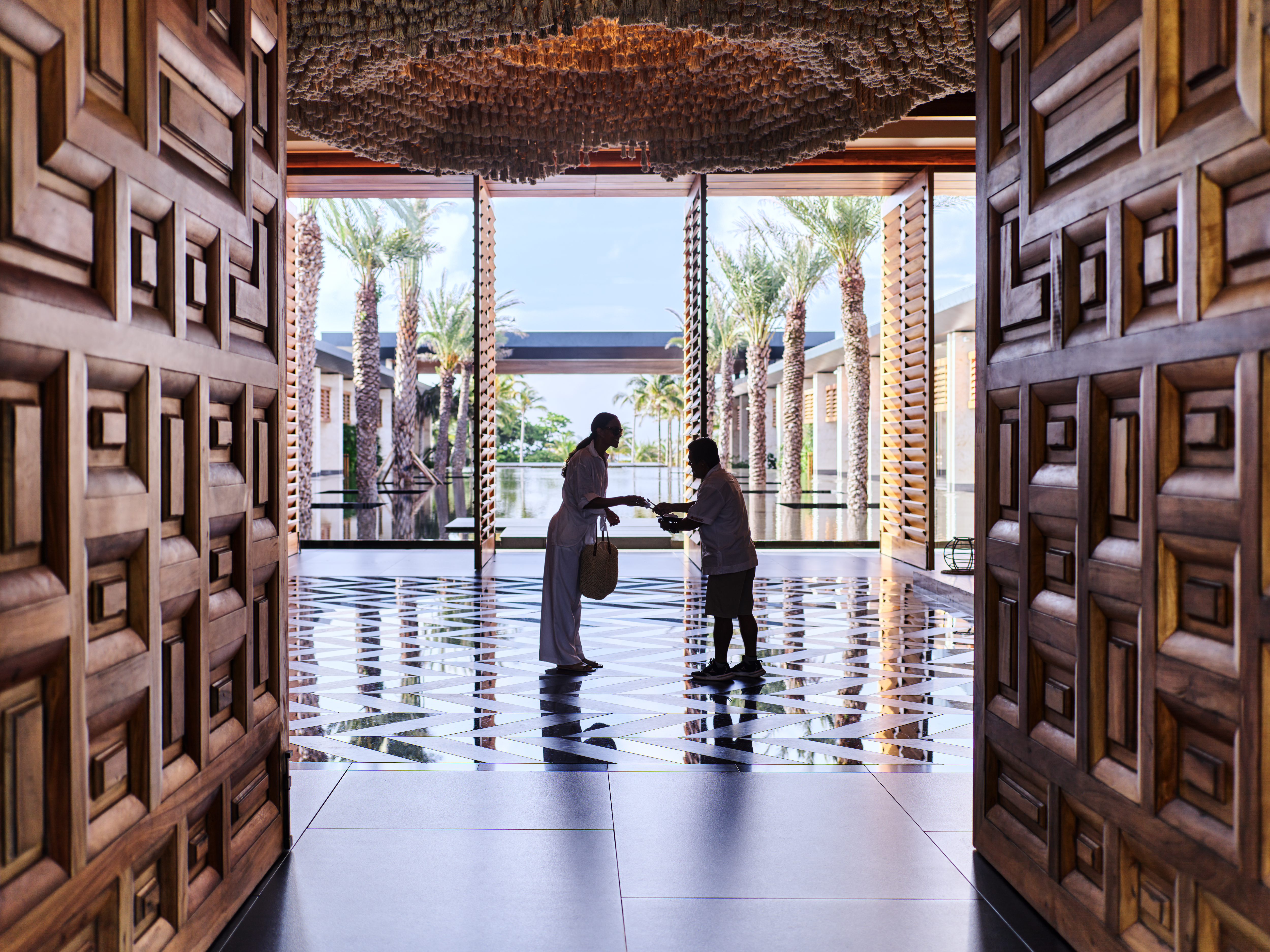 Two people exchanging an item in a spacious, open lobby with large wooden doors, patterned floor, and palm trees outside.