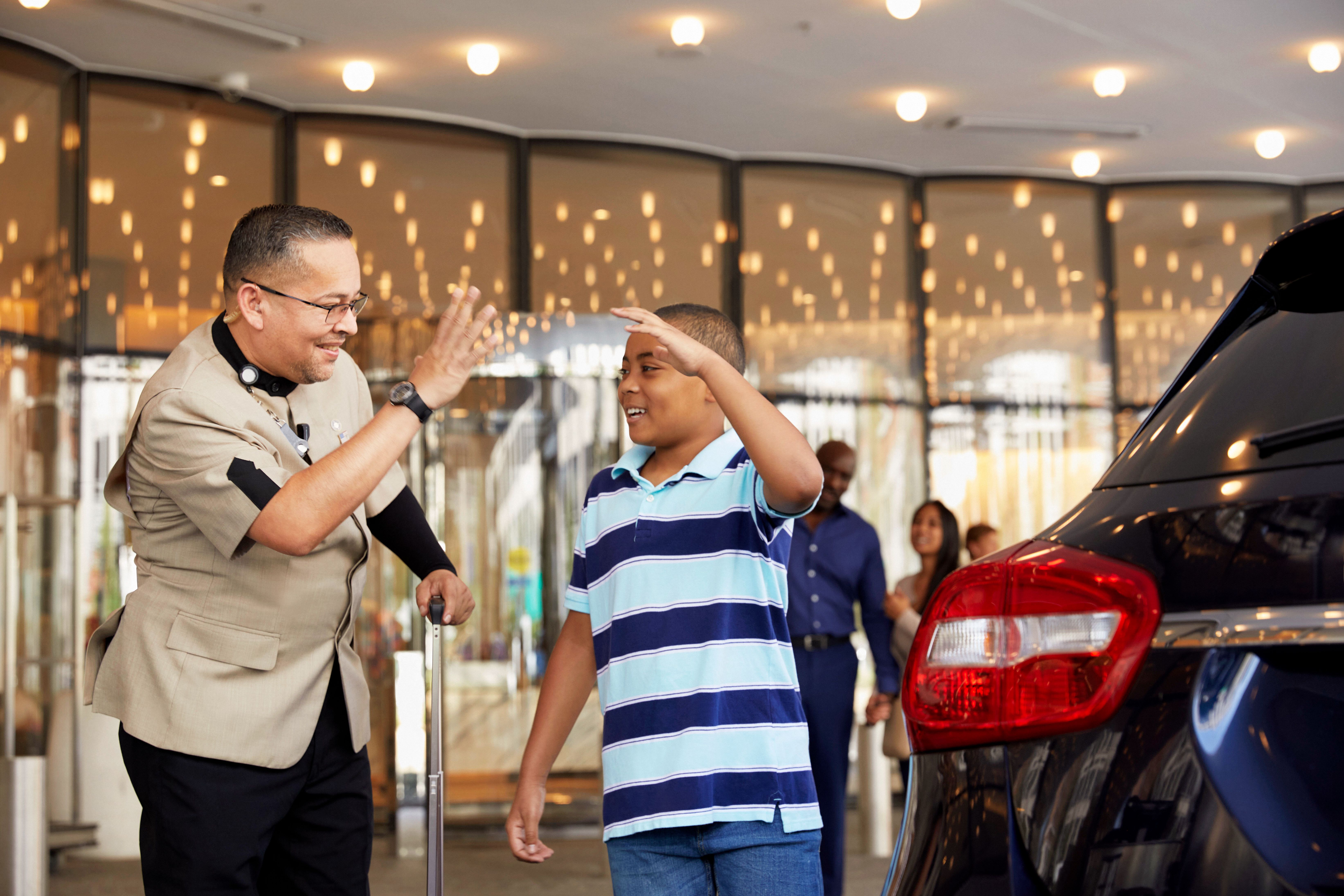 A hotel Bellman and a young guest giving each other a high five outside a building entrance near a parked car.