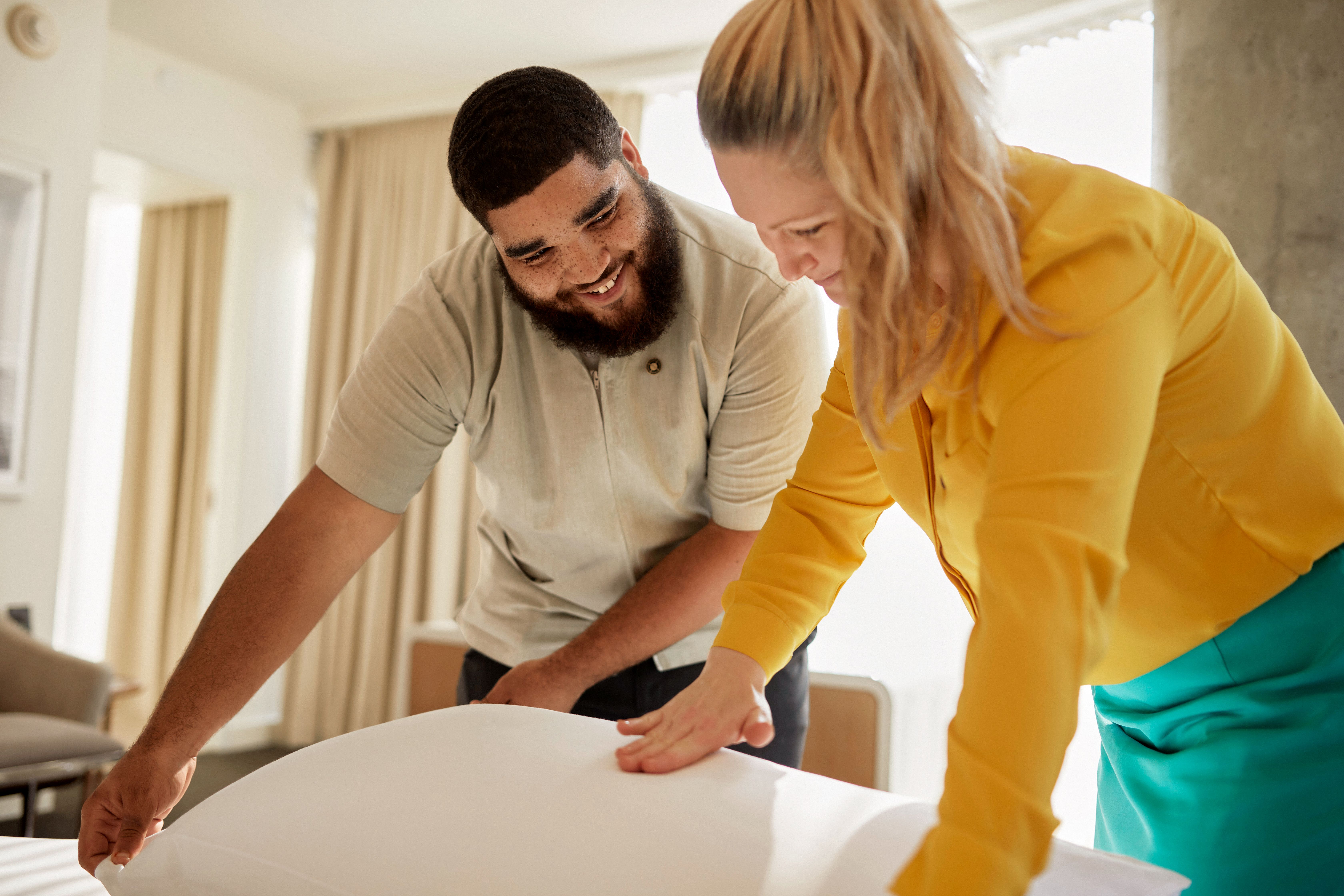 Two people smiling and making a bed together in a bright room.