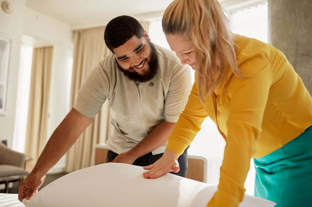 Two people smiling and making a bed together in a bright room.