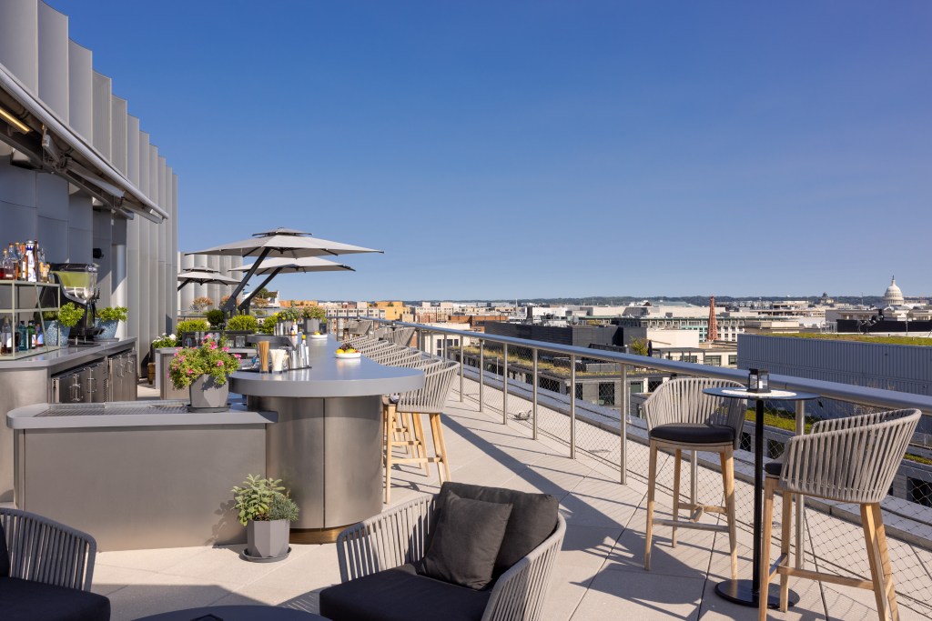 Rooftop bar with modern seating, umbrellas, and a city skyline view under a clear blue sky.