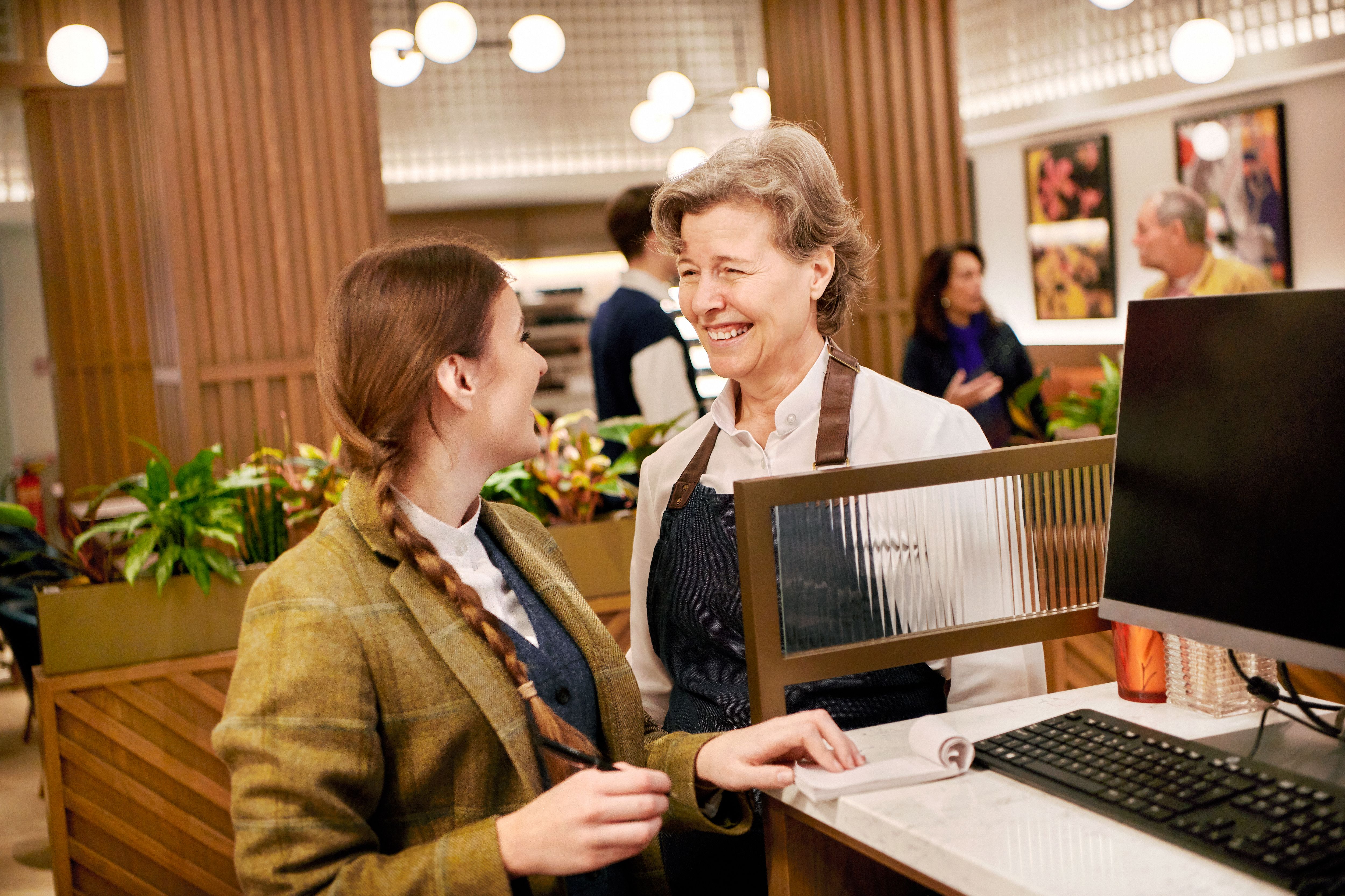 Two women smiling and talking at a reception desk in a warmly lit, modern interior.