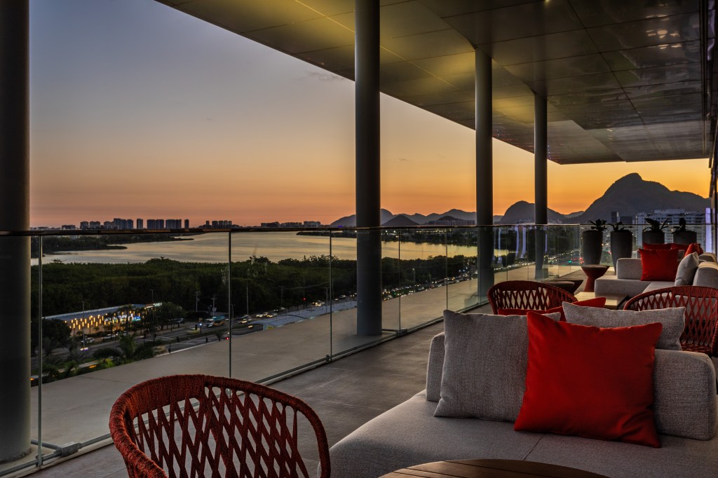 Rooftop balcony seating area with red cushions overlooking water and mountains at sunset.