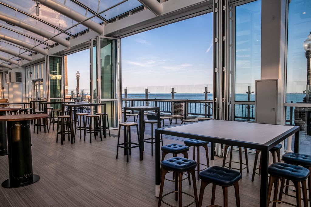 Indoor seating area with high tables and stools overlooking a waterfront through large glass windows.