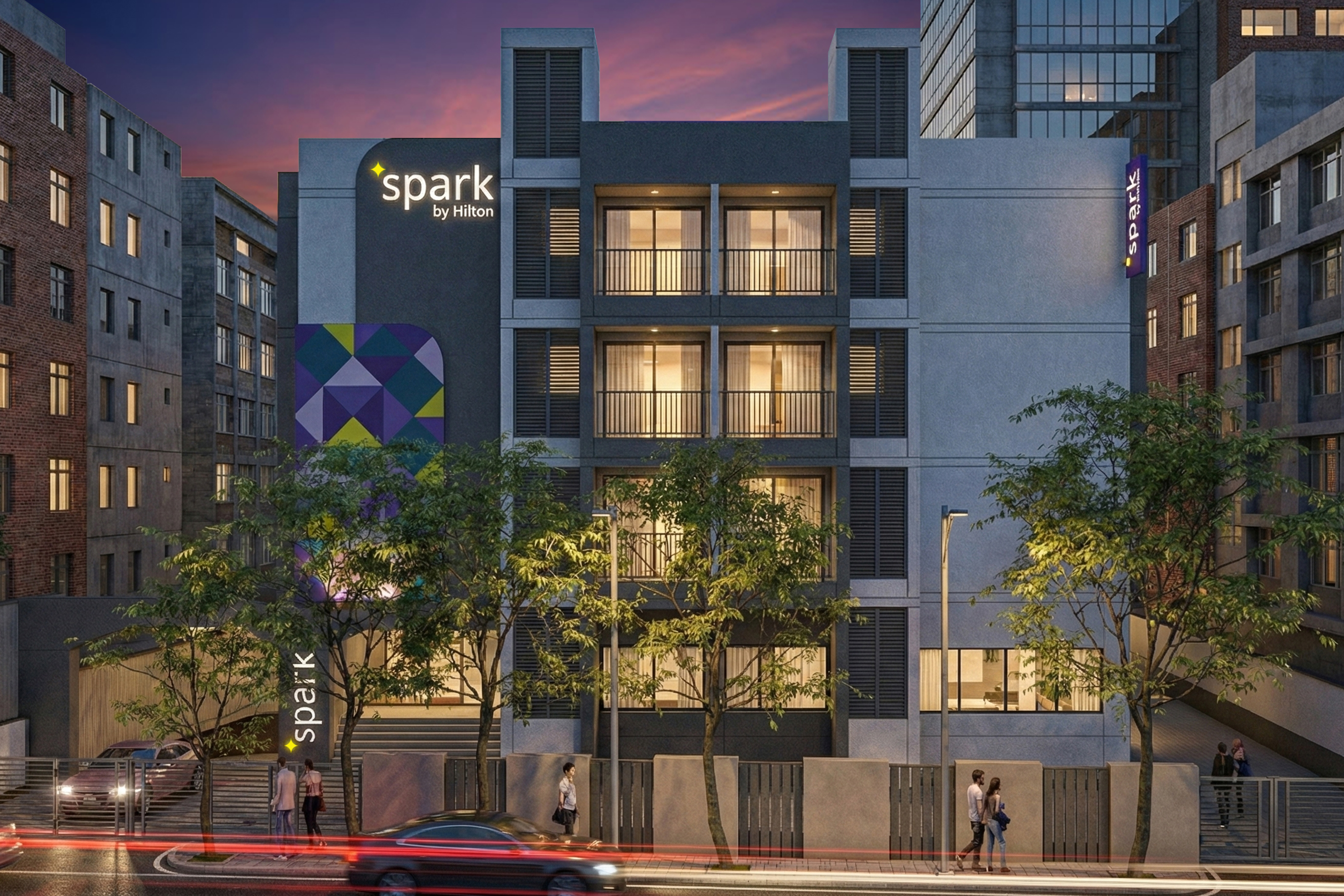 Modern hotel building with illuminated windows and "Spark by Hilton" signage at dusk, surrounded by trees and city buildings.