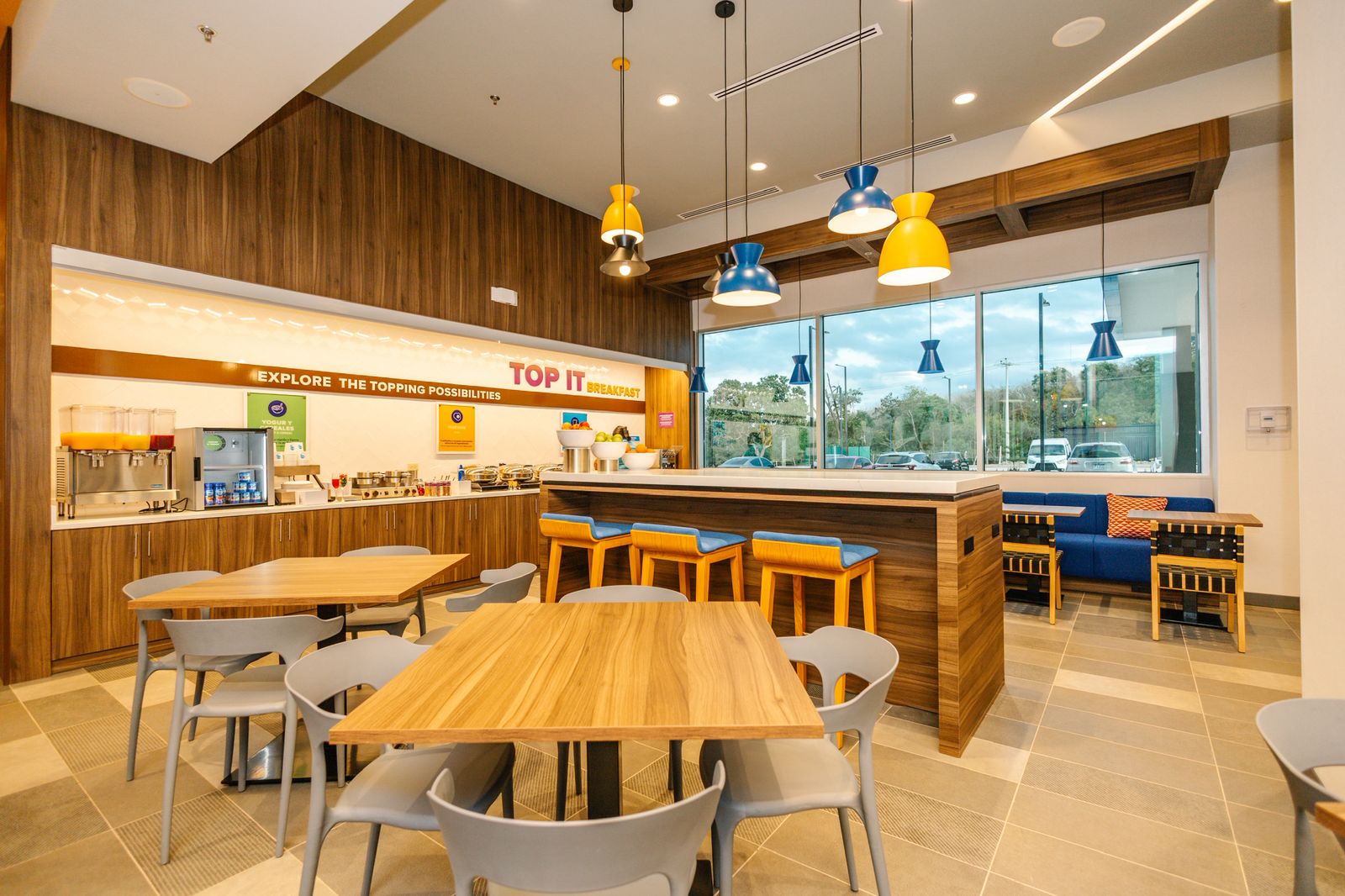 Modern breakfast area with wooden tables, gray chairs, a counter with blue and yellow stools, and a beverage station under warm lighting.