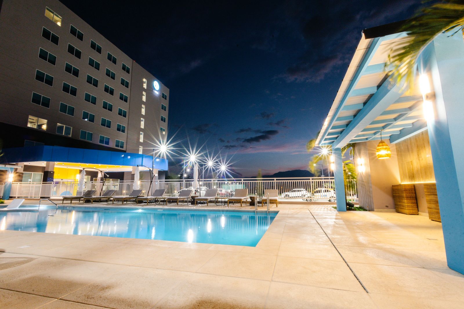 Outdoor swimming pool area at night with lounge chairs, a modern building, and a covered patio with warm lighting.