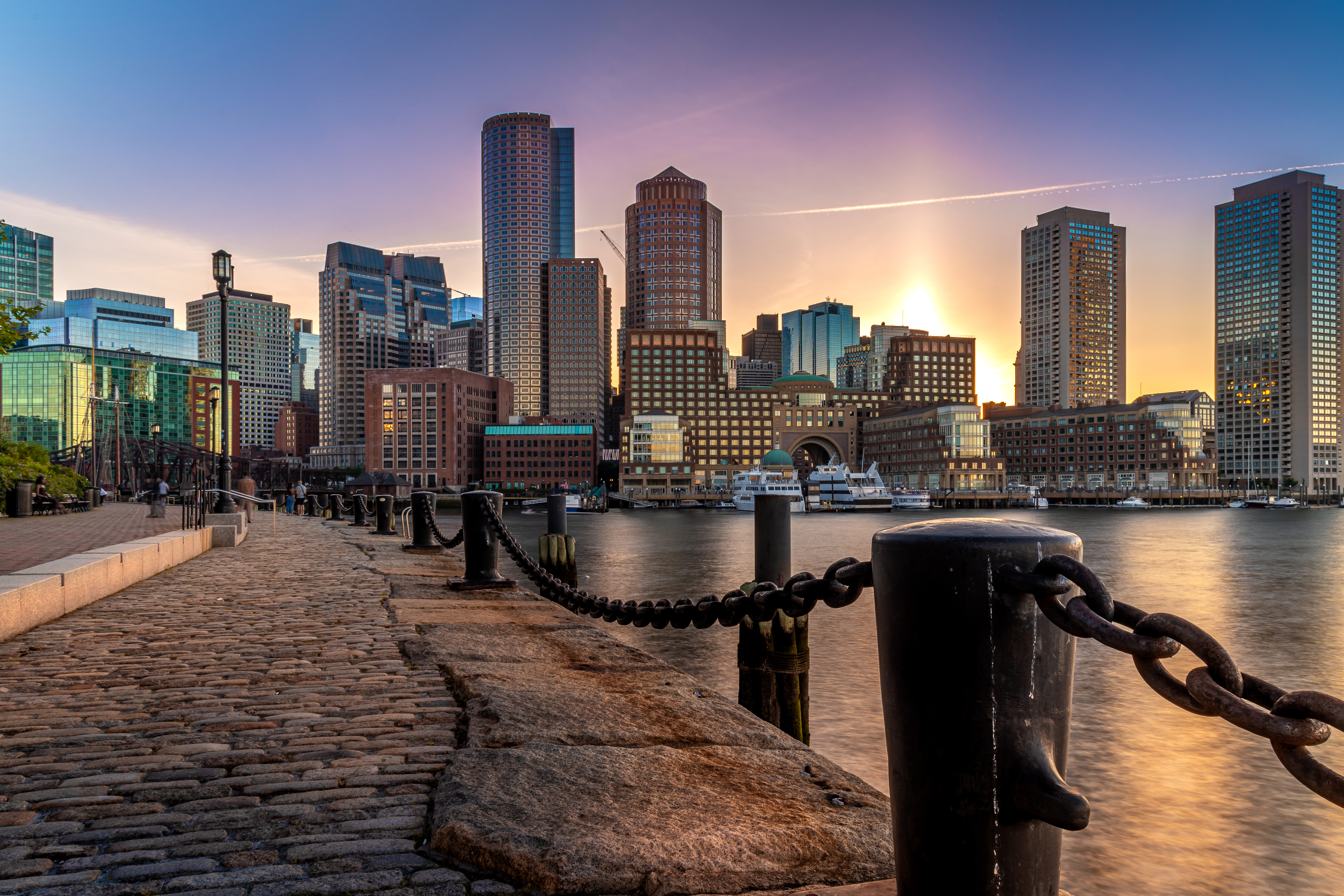 Sunset over a waterfront city skyline with tall buildings and a cobblestone walkway lined with chains and posts.